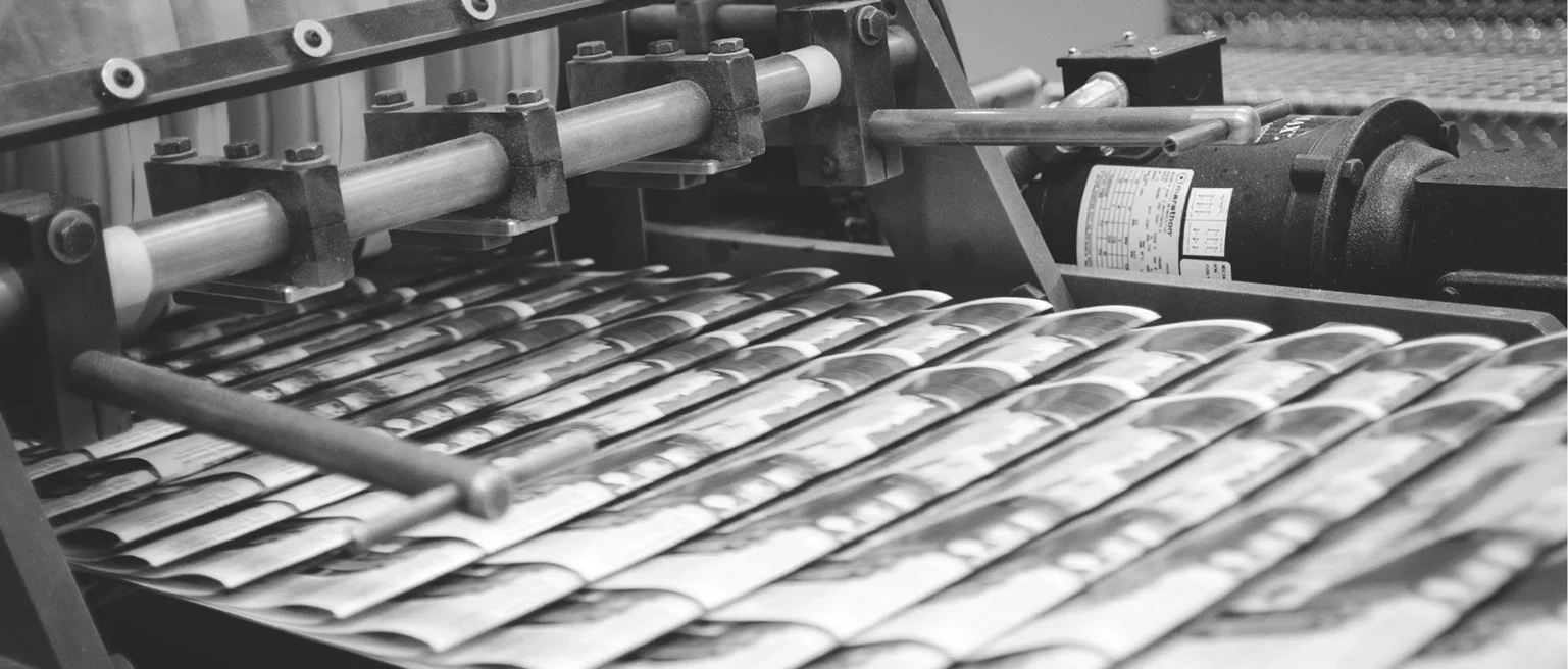 Black and white image of a printing press with papers moving over cylindrical rollers.