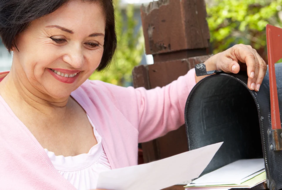A woman smiles while checking her mailbox and holding an envelope.