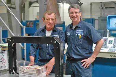 Two men in blue uniforms standing in a printing facility with machinery in the background.